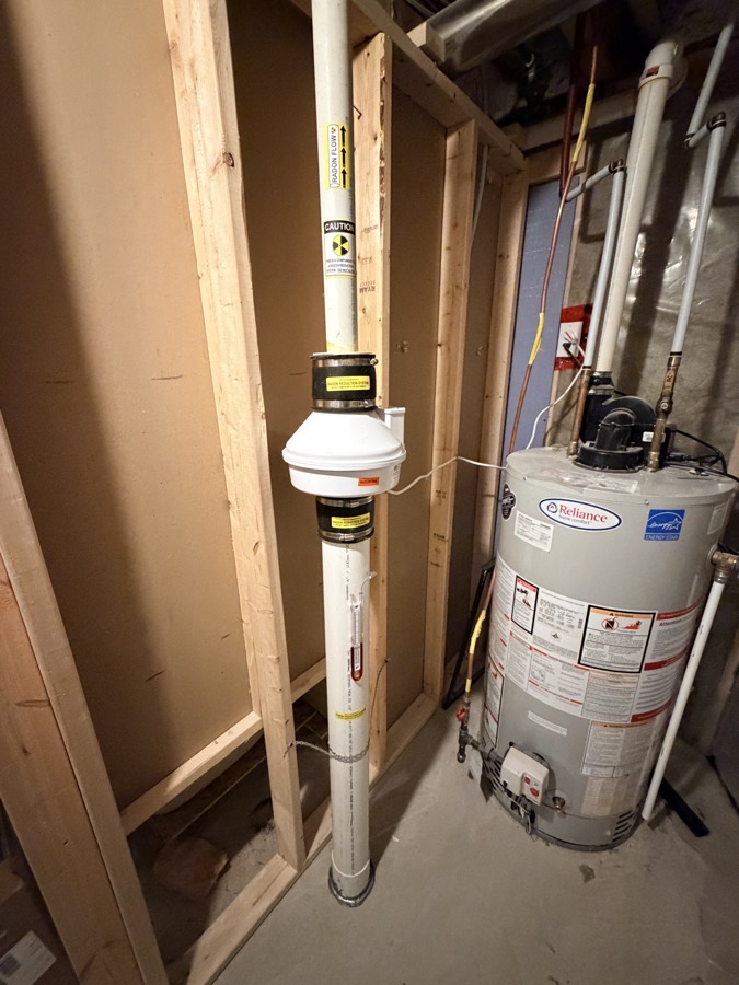 A completed ASD radon mitigation installation in a Toronto basement — vertical exhaust pipe with caution labelling, in-line fan unit, and clean routing alongside the home's water heater